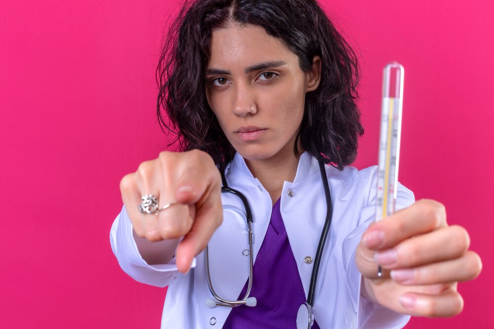 woman doctor wearing white coat with stethoscope showing thermometer pointing displeased to the camera angry and furious with you on isolated pink